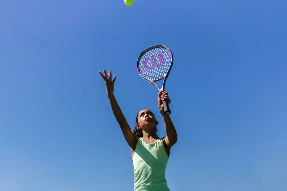 Adolescentă care servește la tenis în aer liber, sub un cer albastru senin